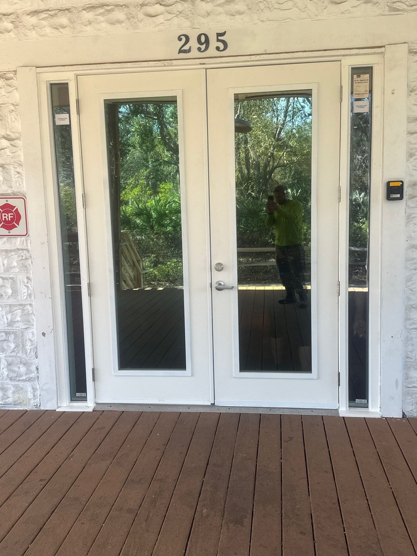 White double entry doors with full-length glass panels and narrow glass sidelites, set into a textured light stone wall above the house number 295, opening onto a brown composite deck