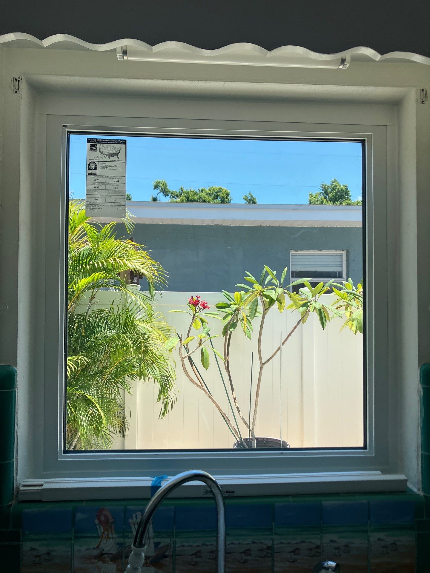 Newly installed impact window above a kitchen sink with the factory energy rating label still on the glass, looking out to a palm and plumeria garden