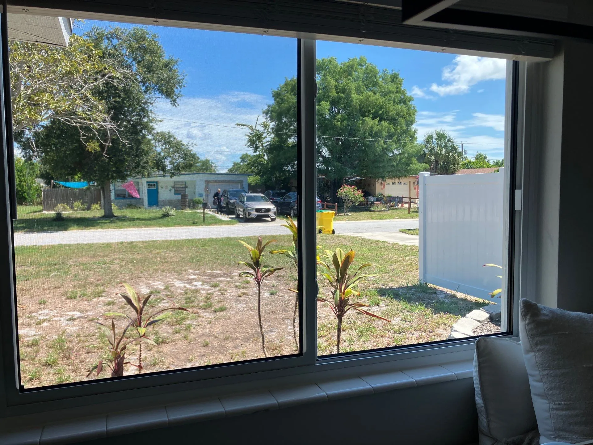 Interior view through a wide sliding window with a deep white sill, looking out to a grassy Florida yard, a white vinyl fence, parked cars, and neighboring homes under a partly cloudy sky