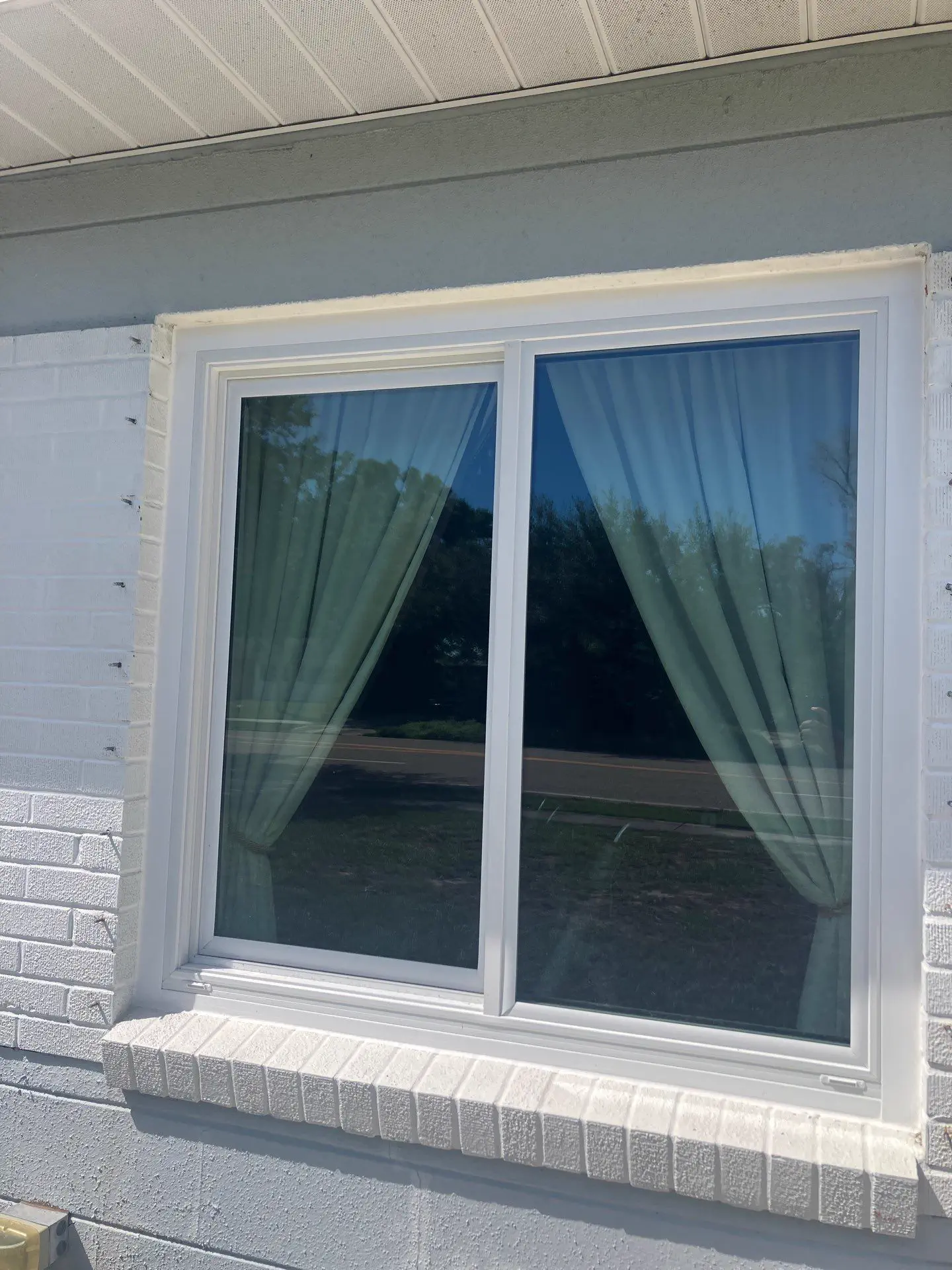 Interior view of an open white single-hung window with the manufacturer's labels still on the glass, showing the metal sill track, hinged sash, and the fenced yard and trees outside.