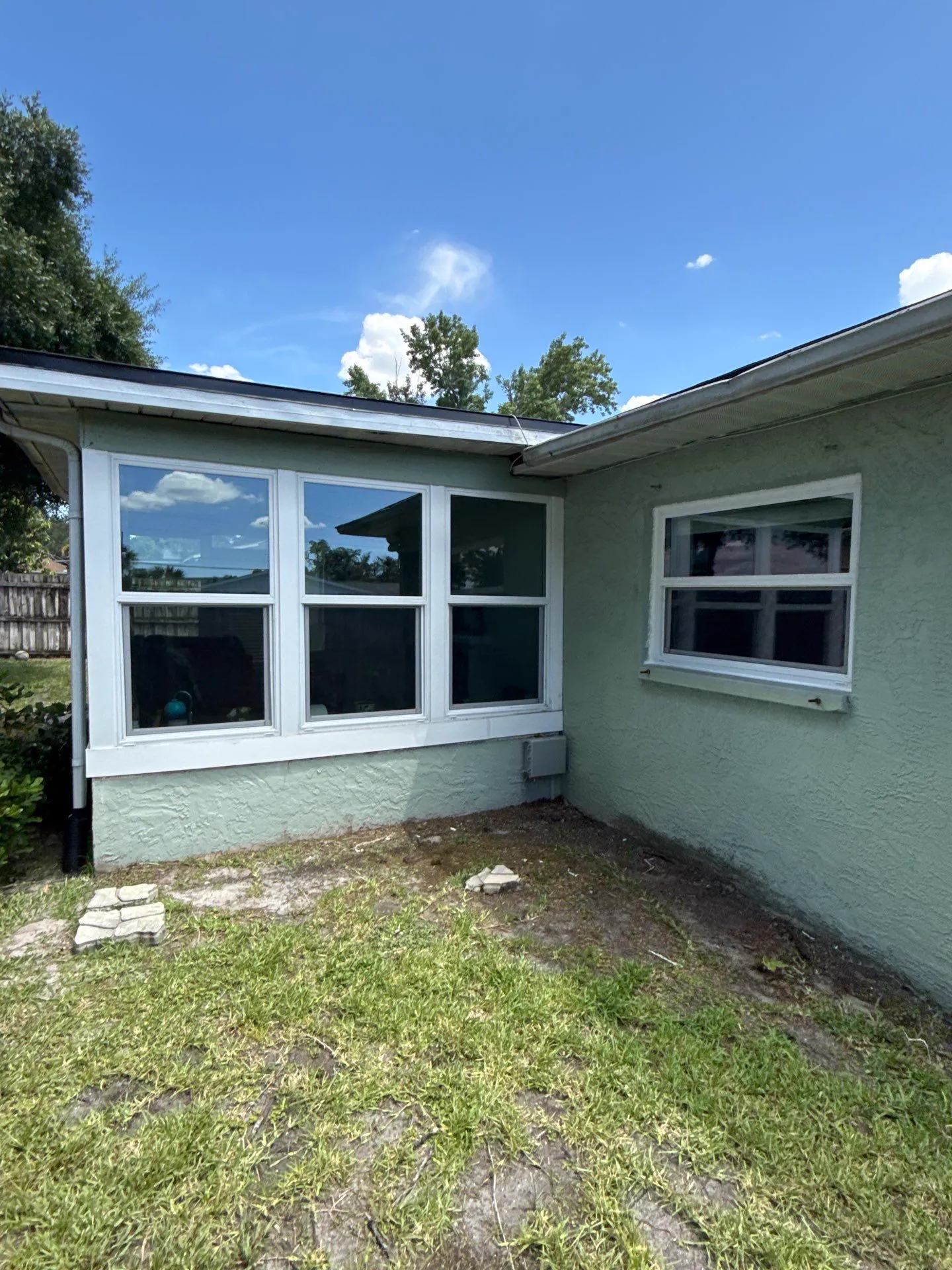 Corner of a light green stucco home where two wings meet, with a row of three white-framed double-hung windows on one wall and a wide white-framed window on the other, under a clear blue sky.