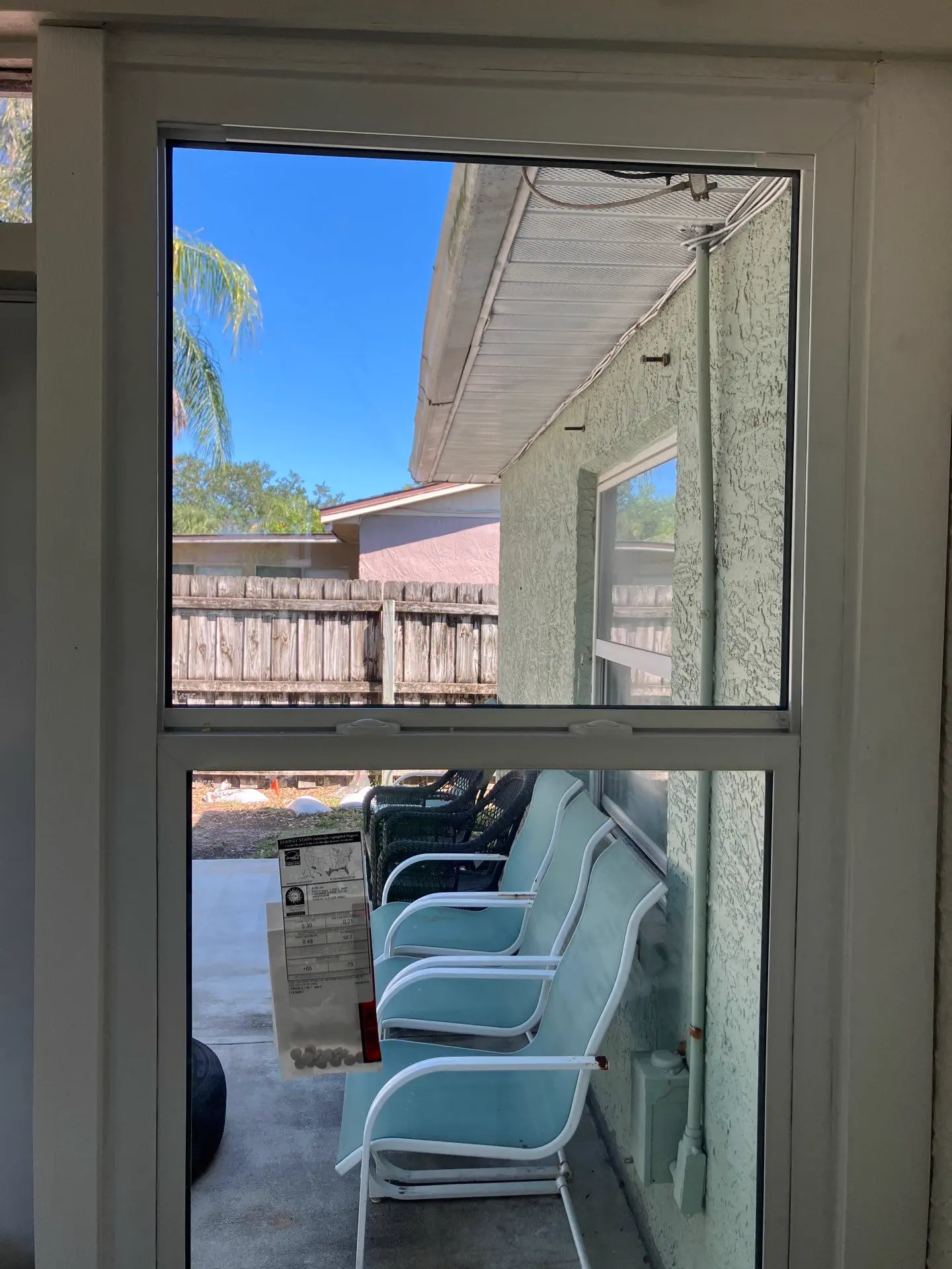 Interior view looking out through a newly installed white single-hung window, with the manufacturer's performance label still attached to the lower sash and a covered patio, stucco wall, and fenced yard visible outside.