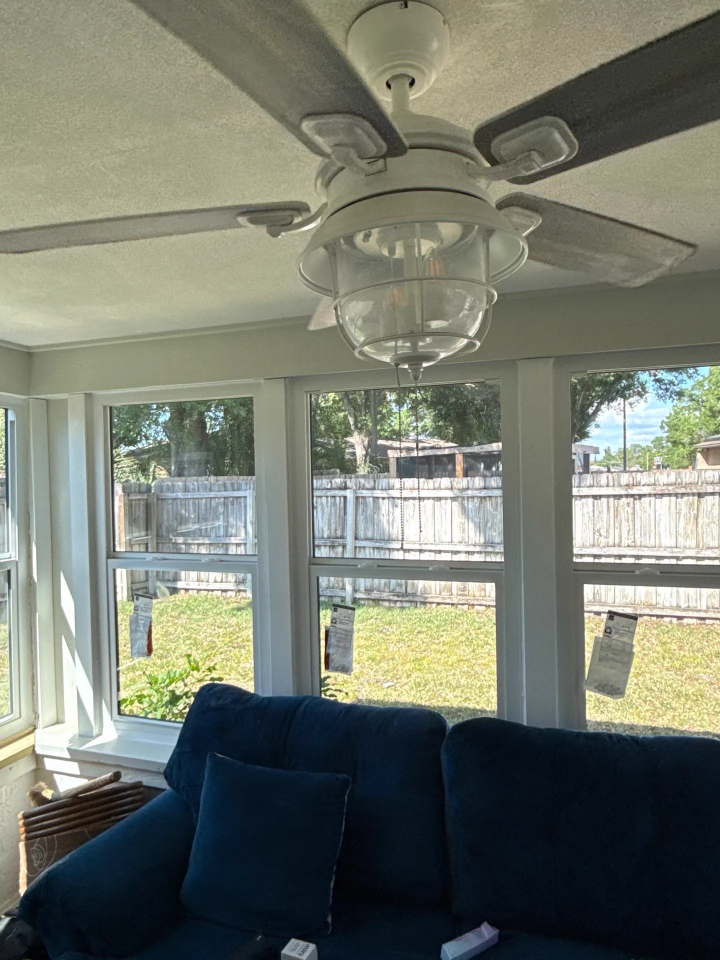 A sunroom with a row of new white-framed windows, product labels still on the glass, looking out to a lawn and a wooden privacy fence, with a white ceiling fan above and a navy sofa below.