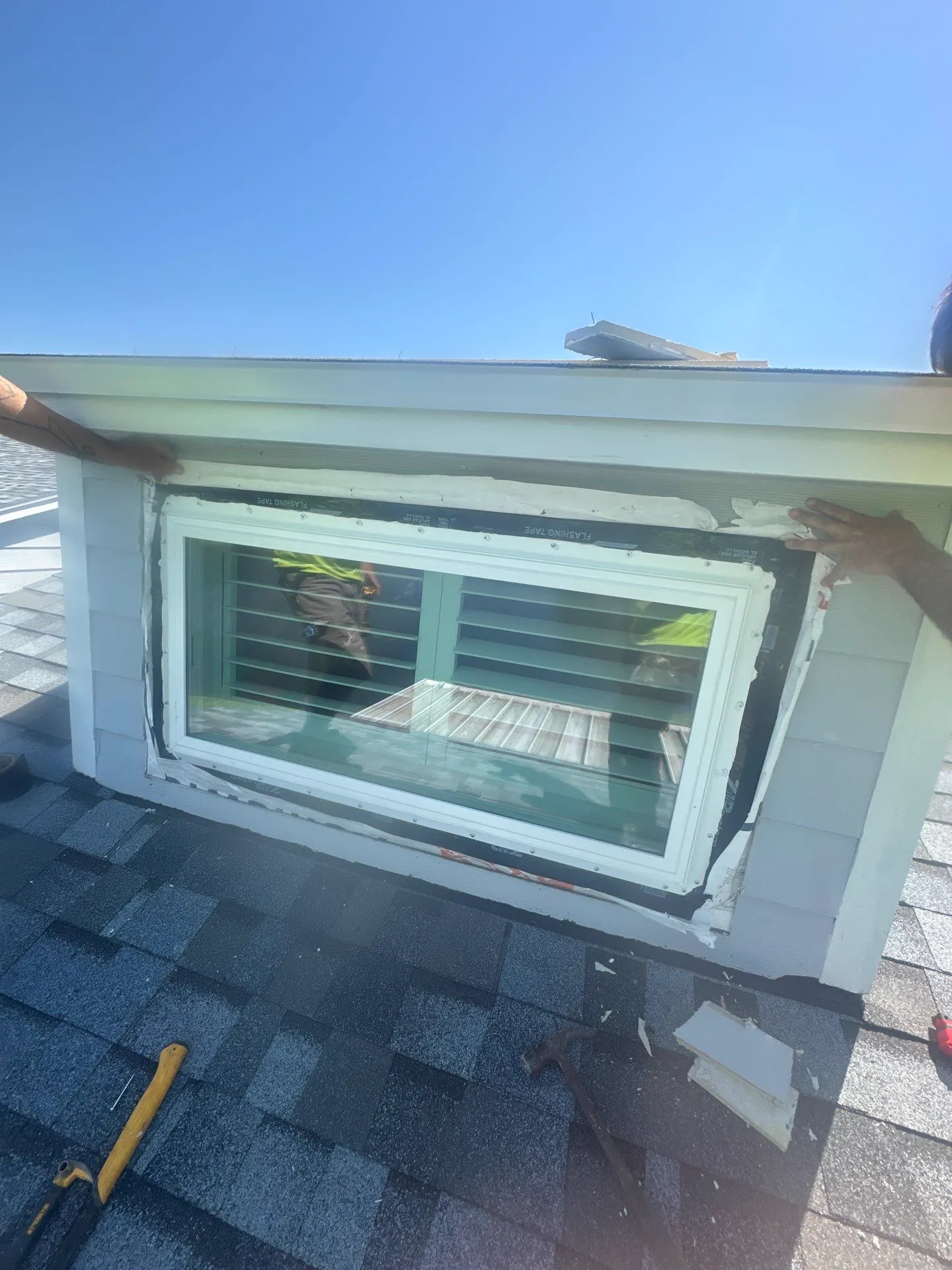 A white-framed dormer window being fitted into an opening on a shingled roof, with the surrounding trim removed and installers' hands at either side of the frame.