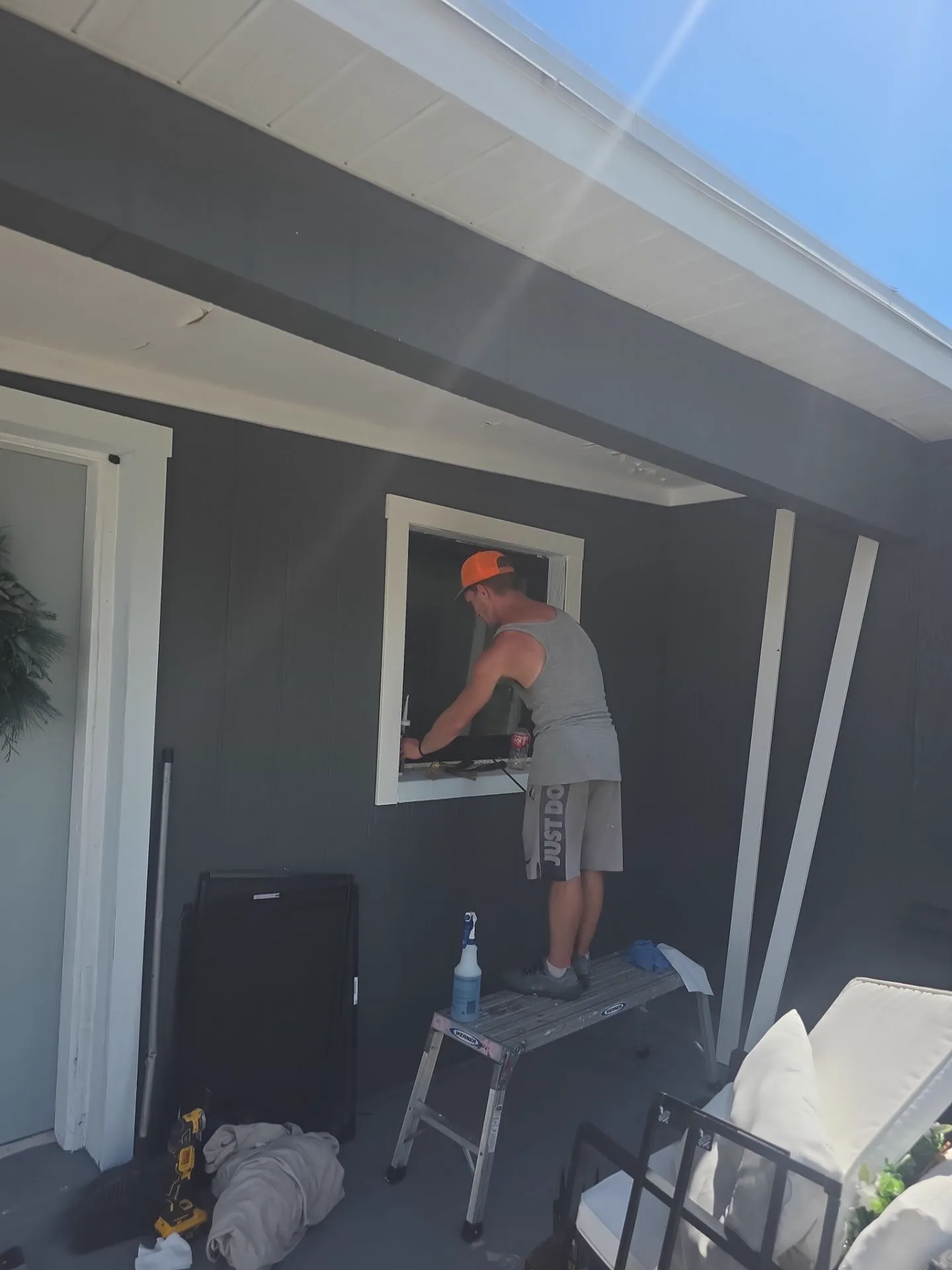 An installer in an orange cap working at a window opening on a covered porch with dark gray siding, standing on a small work platform beside tools and a cleaning bottle.