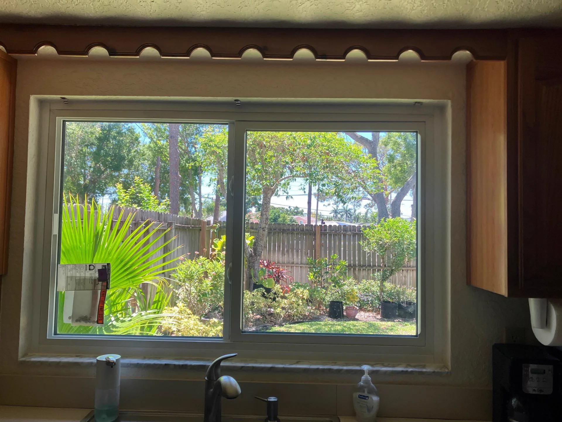 A white-framed sliding window above a kitchen sink, with a product label still on the glass, looking out to palms, garden plants and a wooden fence.
