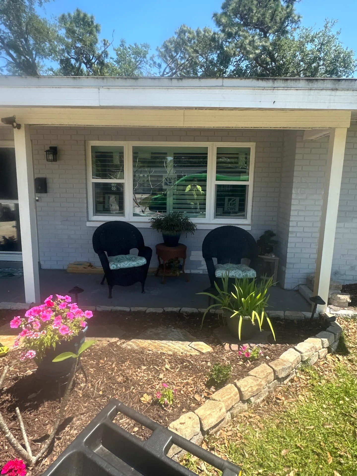Covered front porch of a painted brick home with a wide white-framed window over two wicker chairs, plants and a flower bed in front.
