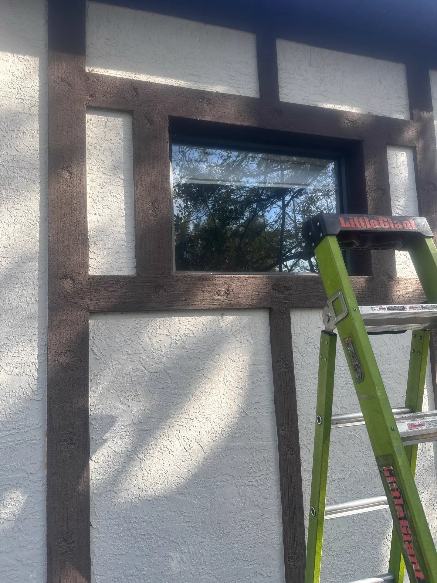 Exterior of a white textured-stucco wall with dark timber trim framing a dark-framed window, with a green Little Giant ladder set up in front of it.
